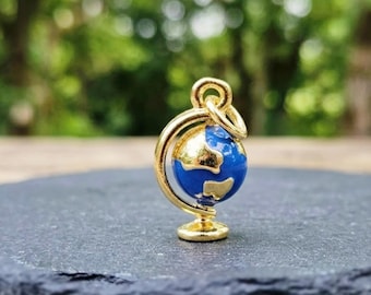 Puede incluir: Un pequeño dije de metal en tono dorado con forma de globo terráqueo, que presenta una esfera de esmalte azul profundo con continentes dorados en relieve y un soporte circular dorado con una anilla superior para colgarlo.
