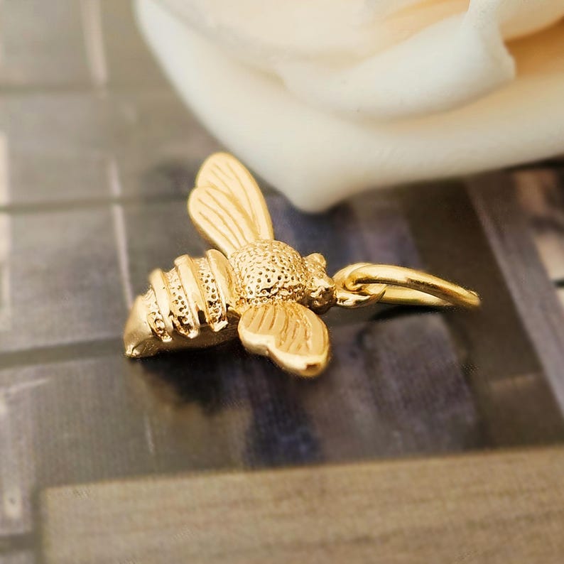 May include: A gold-colored bee charm with detailed wings, body segments, and a small loop for attachment. The bee is positioned at an angle, showcasing its intricate design. The background is blurred, highlighting the charm's details.