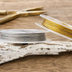 Könnte beinhalten: Zwei durchsichtige Kunststoffspulen, die mit dünnem Metalldraht umwickelt sind, eine mit glänzend goldener und die andere mit silberner Oberfläche, platziert neben einer messingfarbenen Metallzange auf einer strukturierten Oberfläche.