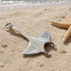 Könnte beinhalten: Ein polierter, silberfarbener Metallanhänger in Form eines Mantarochens mit glatter, reflektierender Oberfläche und einem runden Verbindungsring an der Oberseite. Der Anhänger hat einen breiten, flachen Körper mit spitz zulaufenden Flossen.