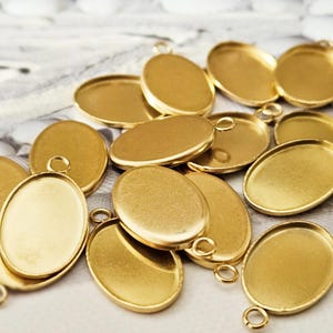Könnte beinhalten: Eine Ansammlung goldfarbener, ovaler Anhänger-Tabletts mit kleinen Schlaufen zum Anbringen von Ketten oder Kordeln. Die Tabletts sind leer, mit einer glatten, glänzenden Oberfläche, und liegen auf einem gemusterten Stoffhintergrund. Diese Schmuckzubehörteile sind ideal für die Herstellung individueller Anhänger.