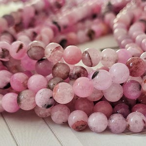 Könnte beinhalten: Eine Ansammlung runder, polierter Perlen in verschiedenen Rosatönen, die von blassem Blush bis zu tiefem Magenta reichen. Jede Perle weist einzigartige, natürlich wirkende dunkelbraune und schwarze Einschlüsse auf, die ein marmoriertes Muster bilden.