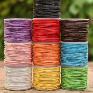 Könnte beinhalten: Eine Sammlung farbenfroher Spulen aus gewachster Baumwollschnur in verschiedenen Farbtönen, darunter Schwarz, Lila, Rot, Braun, Rosa, Orange, Blau, Weiß, Gelb und Grün. Die Schnüre sind ordentlich auf Kunststoffspulen aufgewickelt, ideal für Bastelarbeiten und Schmuckherstellung.