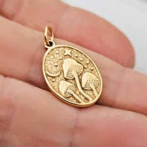 May include: A gold-coloured pendant with an oval shape. The pendant features a design of three mushrooms, a crescent moon, a star, and small dots. The pendant is likely made of metal and has a loop for attaching to a chain.