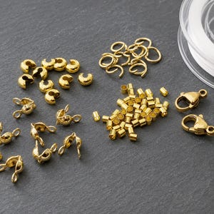 Könnte beinhalten: Eine Auswahl an goldfarbenen Schmuckzubehörteilen, bestehend aus Quetschperlenabdeckungen, Biegeringen, kleinen zylindrischen Quetschperlen und zwei Karabinerverschlüssen, ergänzt durch eine Spule mit transparentem Nylonfaden für Schmuckarbeiten.