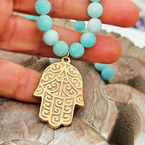 Könnte beinhalten: Eine türkisfarbene Perlenkette mit einem goldfarbenen Hamsa-Hand-Anhänger. Der Anhänger hat aufwendige Schnörkel und hängt an der Kette. Die Perlen sind hellblau.