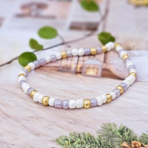 Könnte beinhalten: Ein Perlenarmband mit weißen, lavendelfarbenen und goldfarbenen Perlen. Das Armband ist aufgefädelt und schafft ein flexibles Accessoire. Die Perlen sind in einem Muster angeordnet und sorgen für optische Abwechslung.