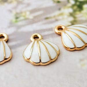 Könnte beinhalten: Nahaufnahme von drei Muschelanhängern. Jeder Anhänger hat ein weißes Emaille-Muscheldesign mit goldfarbenen Akzenten und einer goldfarbenen Schlaufe zum Aufhängen. Die Anhänger sind auf einer weichen, neutral getönten Oberfläche angeordnet.