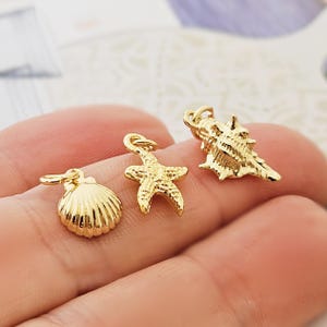 Könnte beinhalten: Drei goldfarbene Anhänger: eine Muschel, ein Seestern und eine Kegelmuschel. Jeder Anhänger hat eine kleine Schlaufe zum Befestigen an einer Halskette oder einem Armband. Die Anhänger sind sehr detailliert und haben ein glänzendes Finish.