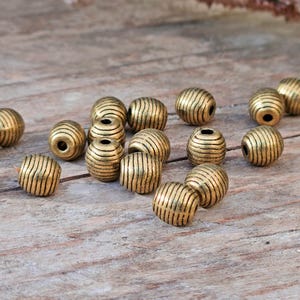 Könnte beinhalten: Eine Sammlung antiker goldfarbener Perlen mit gerilltem, gestreiftem Design. Die Perlen sind auf einer verwitterten Holzoberfläche verstreut und zeigen ihre detaillierte Textur und ihren metallischen Glanz. Ideal für die Schmuckherstellung.