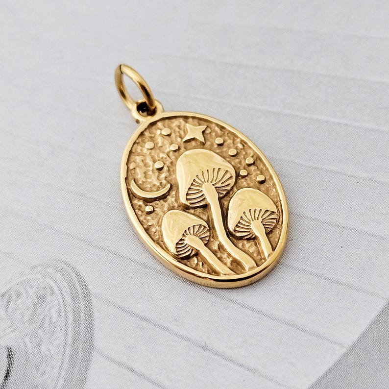 May include: A gold-coloured pendant with an oval shape. The pendant features a design of three mushrooms, a crescent moon, a star, and small dots. The pendant has a loop at the top for a chain.