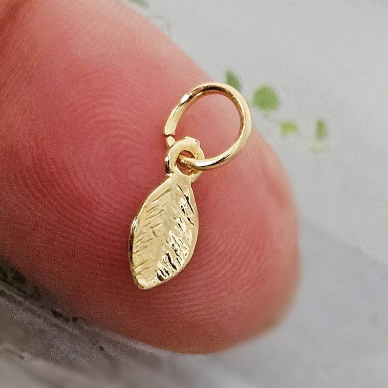 May include: A small, gold-colored leaf charm with a textured surface, attached to a circular ring. The charm is designed to be used as a pendant or for jewelry making. The leaf charm is a delicate and elegant piece.