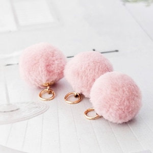 Könnte beinhalten: Drei flauschige, hellrosa Pompons mit goldfarbenen Metallring-Anhängern. Die Pompons sind weich und rund, mit einer flauschigen Textur. Die goldenen Ringe sind an der Basis jedes Pompons befestigt.