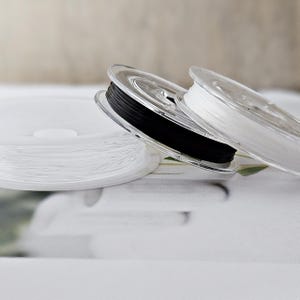 Könnte beinhalten: Nahaufnahme mehrerer Spulen elastischer Schnur in Weiß und Schwarz. Die Schnur ist auf transparenten Kunststoffspulen aufgewickelt. Die Spulen sind in verschiedenen Winkeln angeordnet, wobei sich die weißen Spulen im Vordergrund und die schwarze Spule in der Mitte befinden.