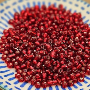 Könnte beinhalten: Nahaufnahme eines Haufens kleiner, durchscheinender roter Glasperlen. Die Perlen sind zylindrisch mit einem Loch in der Mitte. Sie liegen auf einem weißen Teller mit blau-weißem Streifenmuster.