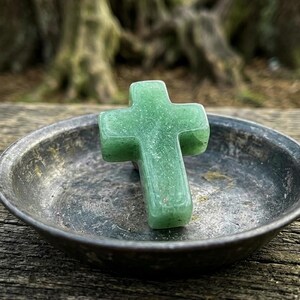 Könnte beinhalten: Ein glattes, hellgrünes Steinkreuz mit gesprenkelter Textur, das in einer flachen, runden Schale aus verwittertem Metall liegt. Das Kreuz hat abgerundete Kanten und eine polierte Oberfläche, die einen Kontrast zur dunklen Metallschale bildet.