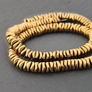 Könnte beinhalten: Nahaufnahme von zwei Strängen goldfarbener, flacher, scheibenförmiger Perlen. Die Perlen sind aneinandergereiht und bilden eine flexible, gebogene Form. Die Perlen haben einen metallischen Glanz und sind vor einem dunkelgrauen Hintergrund angeordnet.