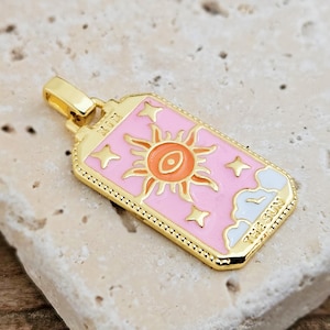 Könnte beinhalten: Goldfarbener Anhänger mit rosa Hintergrund und einem Sonnenmotiv in Orange und Gelb. Der rechteckige Anhänger trägt die weißen Schriftzüge "THE SUN" und "XIX". Das Design ist einer Tarotkarte nachempfunden.