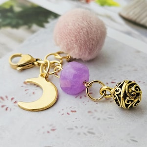 Könnte beinhalten: Ein goldfarbener Schlüsselanhänger mit einem Halbmond-Anhänger, einer facettierten lila Perle und einem filigranen Ball. Er hat auch einen flauschigen, hellrosa Pompon. Der Schlüsselanhänger ist mit einem Karabinerverschluss versehen.