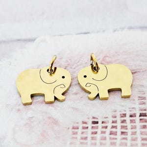 May include: Two gold-colored elephant charms with black details. Each charm has a small loop at the top for attaching to a necklace or bracelet. The charms are on a soft, patterned background.