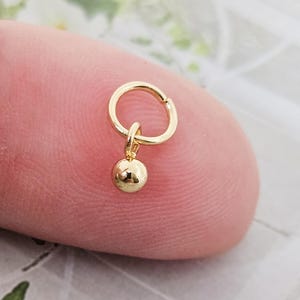 Könnte beinhalten: Ein goldener Creolenohrring mit einem kleinen goldenen Kugelanhänger.