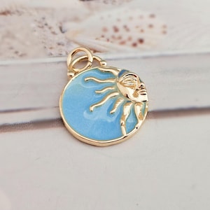 Könnte beinhalten: Ein goldfarbener Anhänger mit einem himmlischen Design. Der Anhänger hat einen hellblauen Emaille-Hintergrund mit einem Sonnengesicht und Strahlen. Das Sonnengesicht ist goldfarben und der Anhänger hat eine Schlaufe zum Befestigen an einer Kette oder einem Armband.