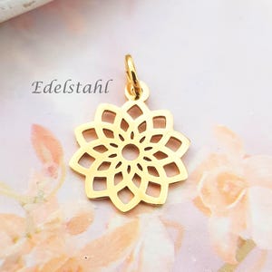 Könnte beinhalten: Goldfarbener Anhänger in Form einer stilisierten Blume mit einem zentralen Loch und durchbrochenen Blütenblättern. Der Anhänger hat oben eine kleine Schlaufe zum Befestigen an einer Kette. Das Wort "Edelstahl" ist auf dem Bild aufgedruckt.