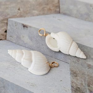 Könnte beinhalten: Zwei weiße Muschelanhänger mit goldfarbenen Drahtschlaufen. Die Muscheln haben eine Spiralform und sind vor einem hellgrauen und blauen Hintergrund platziert. Diese Anhänger sind ideal für die Herstellung von Schmuck mit Strandmotiven.