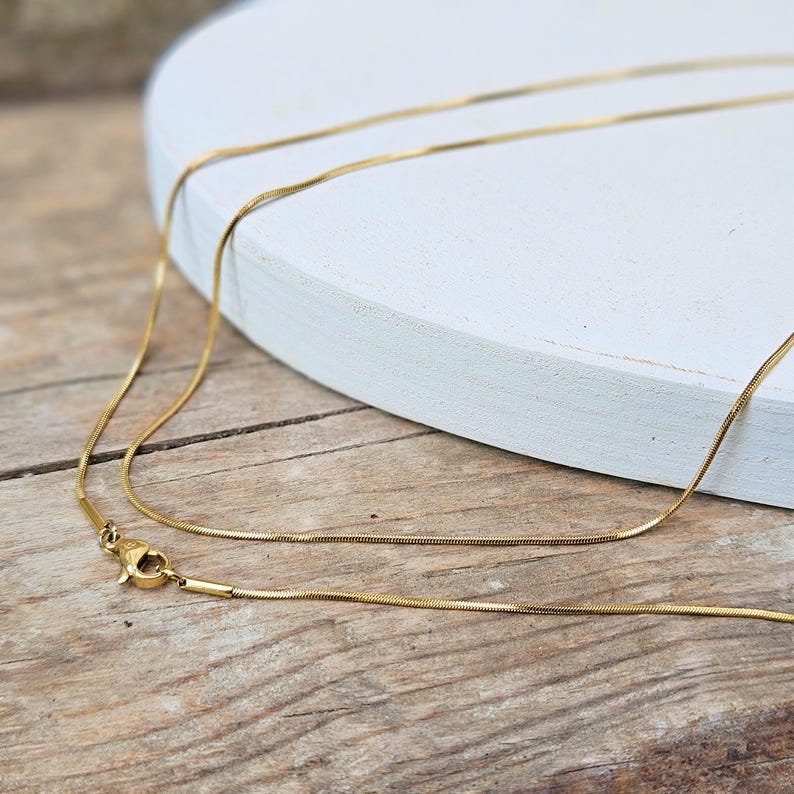 May include: A gold chain necklace with a delicate, snake-like design. The chain is layered on a white surface.
