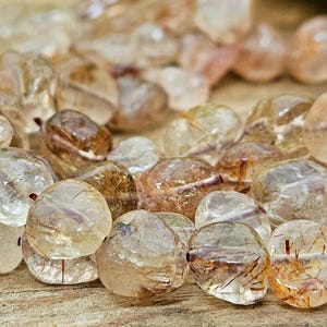 Könnte beinhalten: Nahaufnahme einer Sammlung polierter, klarer Quarzperlen mit goldenen Rutil-Einschlüssen. Die Perlen variieren in Größe und Form, mit einer Mischung aus transparenten und undurchsichtigen Bereichen. Die Farbpalette ist eine warme Mischung aus Gold, Braun und Klar.