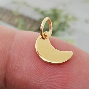 May include: A gold-colored crescent moon charm with a small loop for attaching to a necklace or bracelet. The charm is smooth and shiny, reflecting light. The moon shape is a classic symbol.