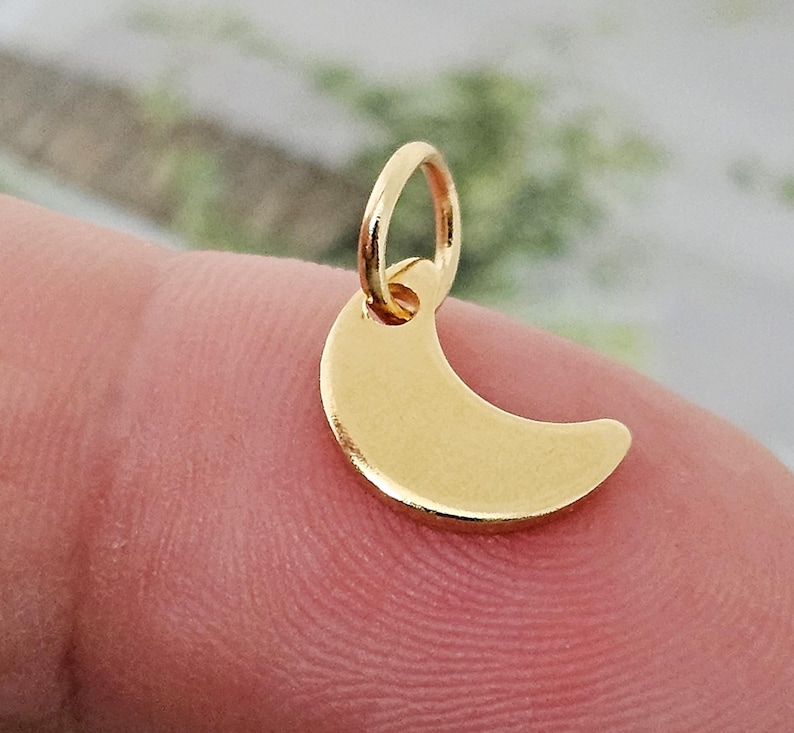 May include: A gold-colored crescent moon charm with a small loop for attaching to a necklace or bracelet. The charm is smooth and shiny, reflecting light. The moon shape is a classic symbol.