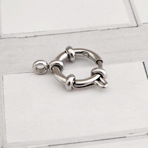 Könnte beinhalten: Ein silberfarbener, runder Federringverschluss. Der Verschluss hat ein kreisförmiges Design mit einer kleinen Schlaufe auf der einen Seite und einem Hebelverschluss auf der anderen Seite. Das Metall hat eine glänzende Oberfläche. Durchmesser ca. 1,5 cm.