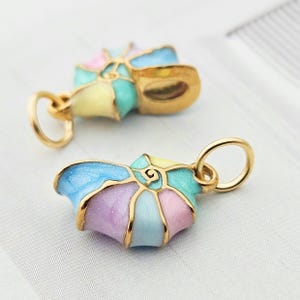 Könnte beinhalten: Zwei bunte Emaille-Muschel-Anhänger mit goldfarbenen Metallakzenten. Die Anhänger weisen Pastellfarben wie Blau, Rosa, Gelb und Lila auf. Jeder Anhänger hat eine kleine Schlaufe zum Befestigen an einer Halskette oder einem Armband.