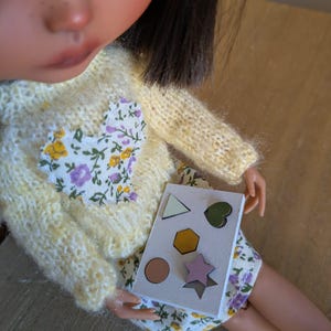 Puede incluir: Una tarjeta blanca con cinco formas de diferentes colores. Las formas son un triángulo, un corazón, un hexágono, un círculo y una estrella. La tarjeta está siendo sostenida por una muñeca.