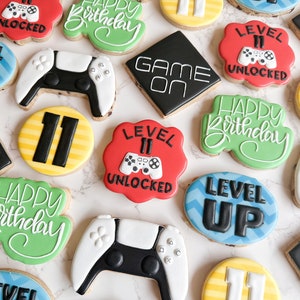 May include: A collection of decorated sugar cookies featuring a video game theme. The cookies are decorated with various designs, including a game controller, the words "Level Up", "Game On", and "Happy Birthday", and the number "11".