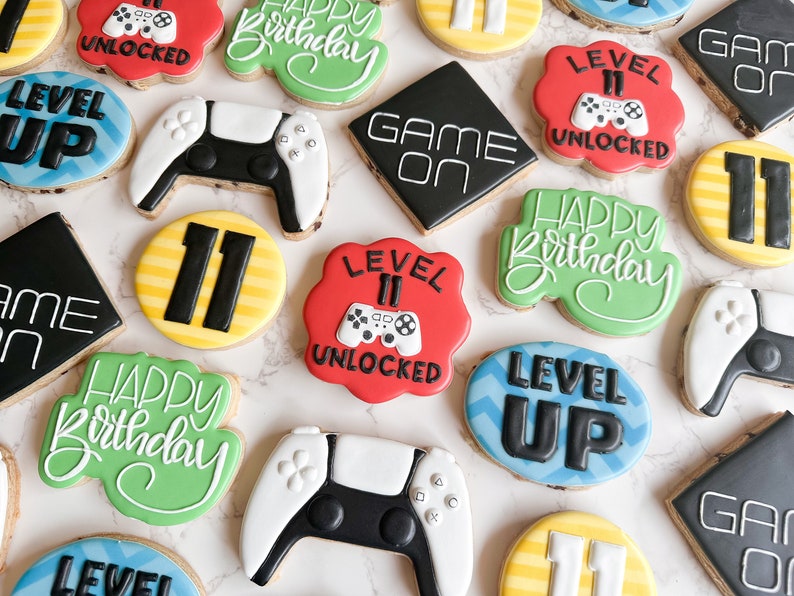 May include: A collection of decorated sugar cookies featuring a video game theme. The cookies are decorated with various designs, including a game controller, the words "Level Up", "Game On", and "Happy Birthday", and the number "11".