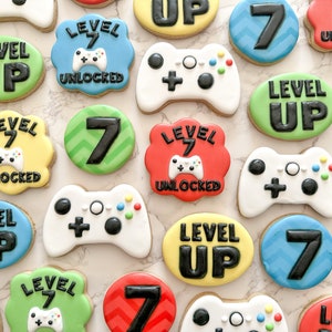 May include: A collection of decorated sugar cookies with a video game theme. The cookies feature various designs, including controllers, the number 7, and the phrase "Level Up".