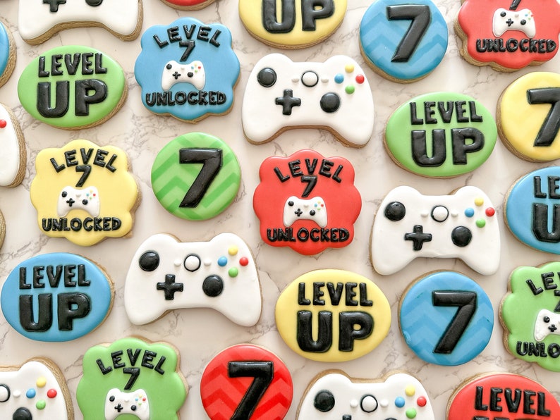 May include: A collection of decorated sugar cookies with a video game theme. The cookies feature various designs, including controllers, the number 7, and the phrase "Level Up".