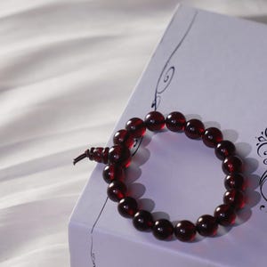 May include: A deep red beaded bracelet lies on a white box with the name "Anna" printed on it. The bracelet is made of round, polished beads and is displayed on a white, textured surface.