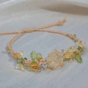 May include: A delicate bracelet made with natural stones, featuring a large, clear quartz crystal surrounded by smaller, yellow and green stones. The bracelet is tied with a brown cord and has a floral design.
