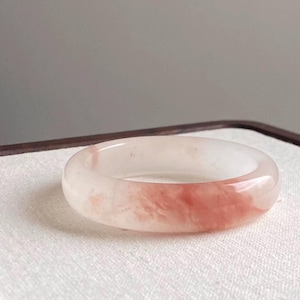 Puede incluir: Una pulsera redonda translúcida con una mezcla de tonos blancos y rosas. La pulsera tiene una superficie lisa y pulida y un aspecto veteado. La pulsera de piedras preciosas es una joya.