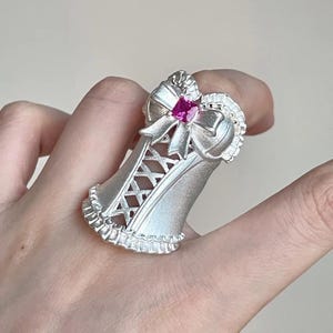 May include: A silver-toned ring shaped like a corset with a matte finish, featuring a central pink square-cut gemstone at the top, a bow detail, and delicate ruffled edges along the bottom and sides, with a crisscross lace-up pattern on the front.