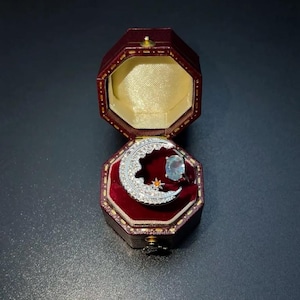 May include: A silver-toned ring featuring a crescent moon motif encrusted with clear stones, a small orange star accent, and a single oval light blue gemstone, presented inside an open, octagonal, dark red velvet-lined jewelry box.