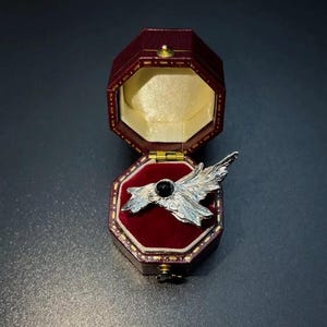 May include: A silver-toned brooch shaped like a bird with outstretched wings, featuring a small, round, black stone at its center. The brooch is displayed inside an open, octagonal, dark red velvet-lined jewelry box with gold-colored trim.