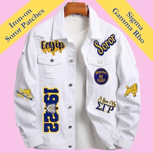 May include: White denim jacket with distressed details, adorned with iron-on patches. Patches include text and graphics in yellow and blue, such as "Eeyip", "Soror", "1922", and "I love My ΣΓΡ". The jacket is hanging on a wooden hanger.