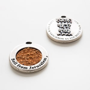 May include: Two silver round pendants. One pendant has a small sample of soil from Jerusalem inside a clear window. The other pendant has Hebrew text that reads "Love your neighbor as yourself."