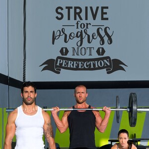 Puede incluir: Dos personas levantando pesas en un gimnasio. El texto en la pared dice "STRIVE for progress NOT PERFECTION".
