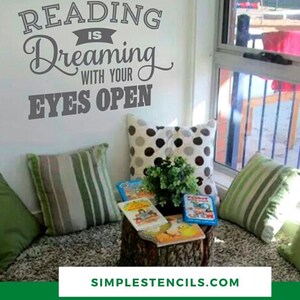 May include: A cozy reading nook with decorative pillows, books, and a small plant on a tree stump. The wall features a gray decal that reads "Reading is Dreaming with your Eyes Open".