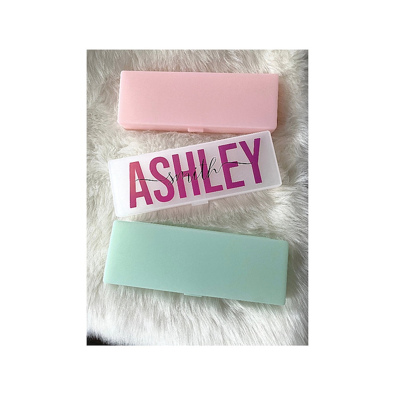 May include: Three rectangular plastic boxes with lids. The middle box is clear with the name "ASHLEY smith" printed in pink letters. The top box is pink and the bottom box is light green.